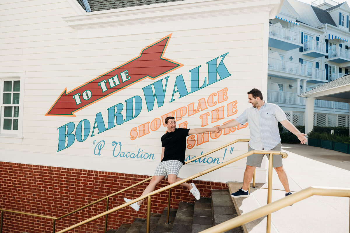 One groom playfully pulls the other down the steps in front of the “Boardwalk” mural at Disney’s Boardwalk resort area.