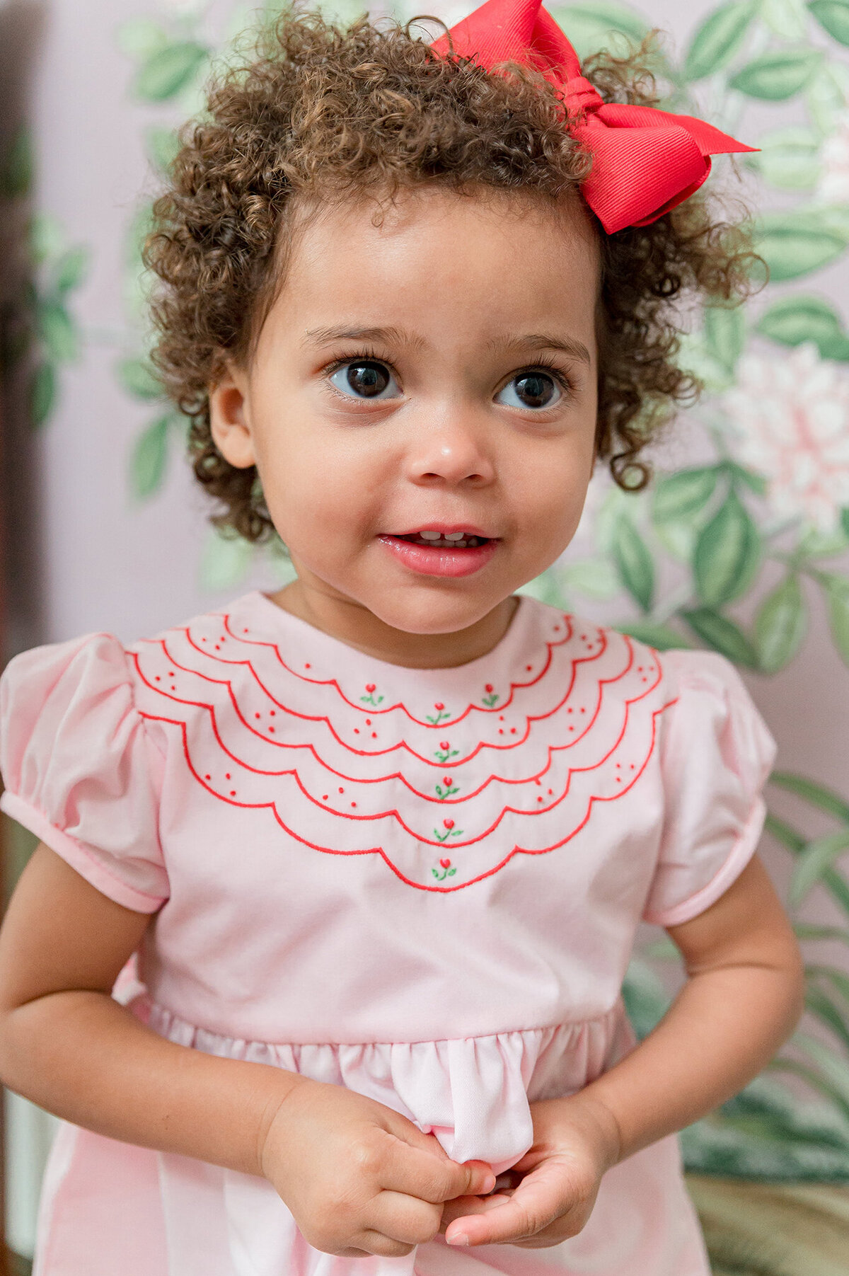 Curly headed girl wearing a pink romper with scalloped red embroidery around the neck.
