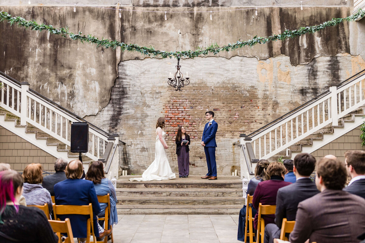 A bride and groom stand with an officiant on a small stage between two staircases, exchanging vows outdoors. Guests are seated, watching. The backdrop is a weathered brick wall with a chandelier overhead.