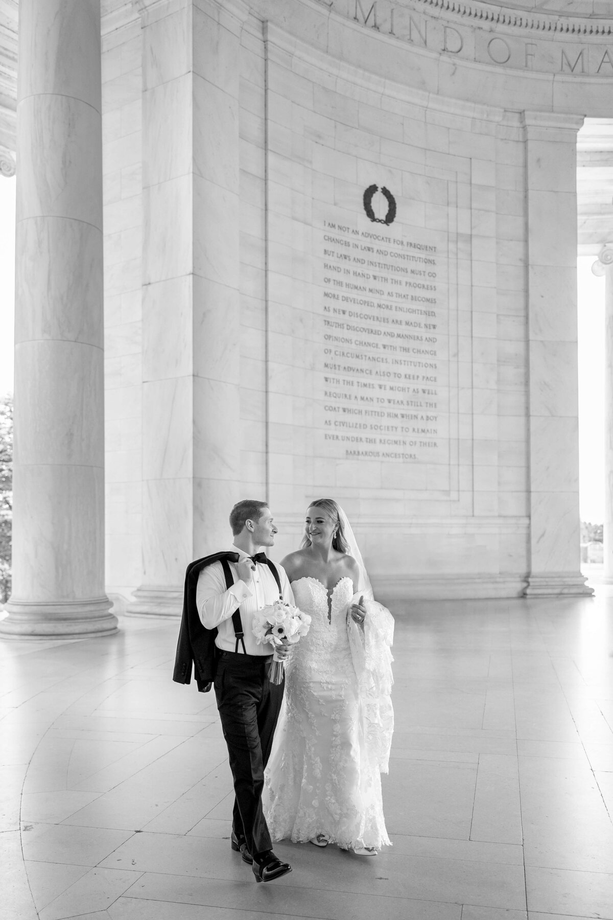 A newlywed couple walks inside a historic building with tall marble columns. The groom holds his suit jacket over his shoulder, while the bride carries a bouquet. Both look at each other with smiles. The backdrop includes engraved text on the wall.