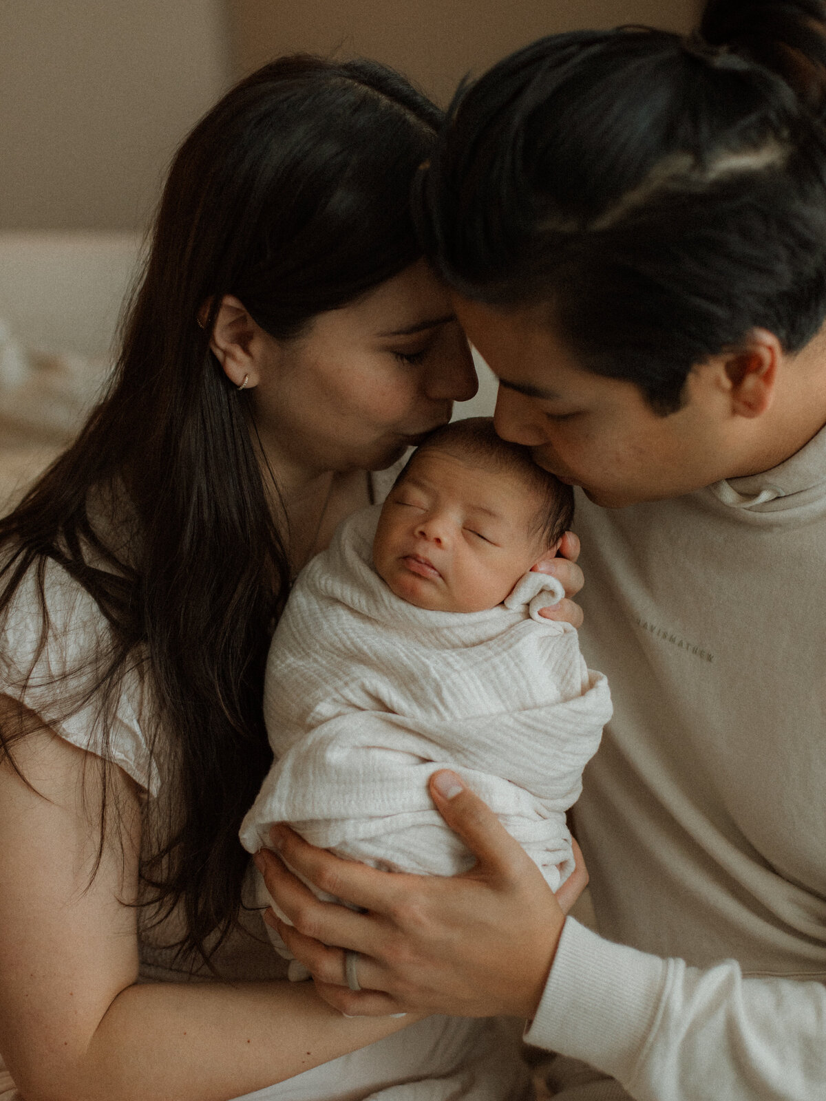 A tender moment as both parents gently kiss their baby’s head, wrapped in a white swaddle.