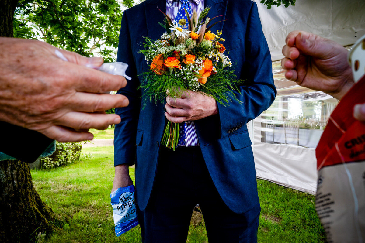A photography of a wedding guest holding a bouquet whilst the other hand he has pipers crisps. his head is cropped off to add a illusion of anonymity