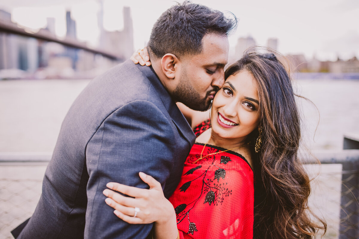 Groom kissing the bride wearing indian sari during their engagement session at the brooklyn bridge park in dumbo