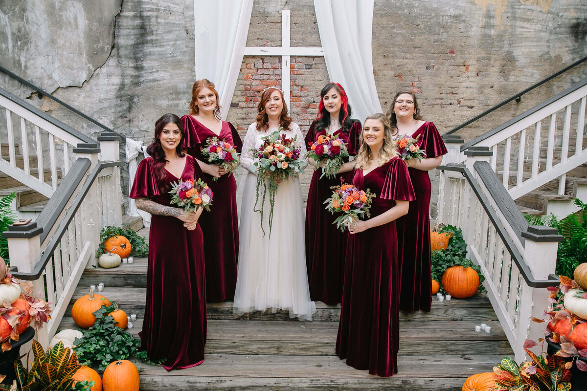 A bride in a white dress stands with five bridesmaids in matching burgundy dresses, all holding bouquets, on wooden stairs decorated with pumpkins and greenery. A white cross and drapes are displayed behind them.