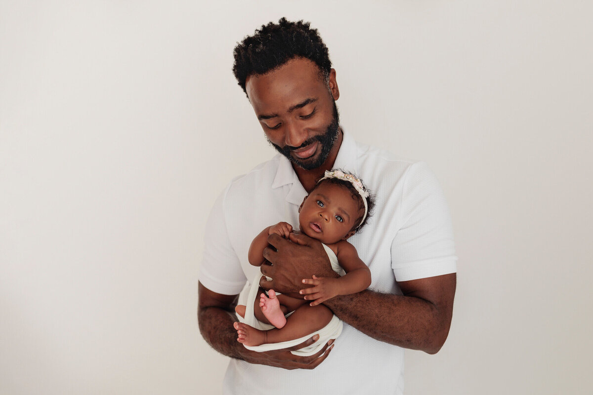 Smiling father in a white shirt holding his newborn baby girl, who is looking at the camera, while wearing a white outfit and a white headband, against a white backdrop.