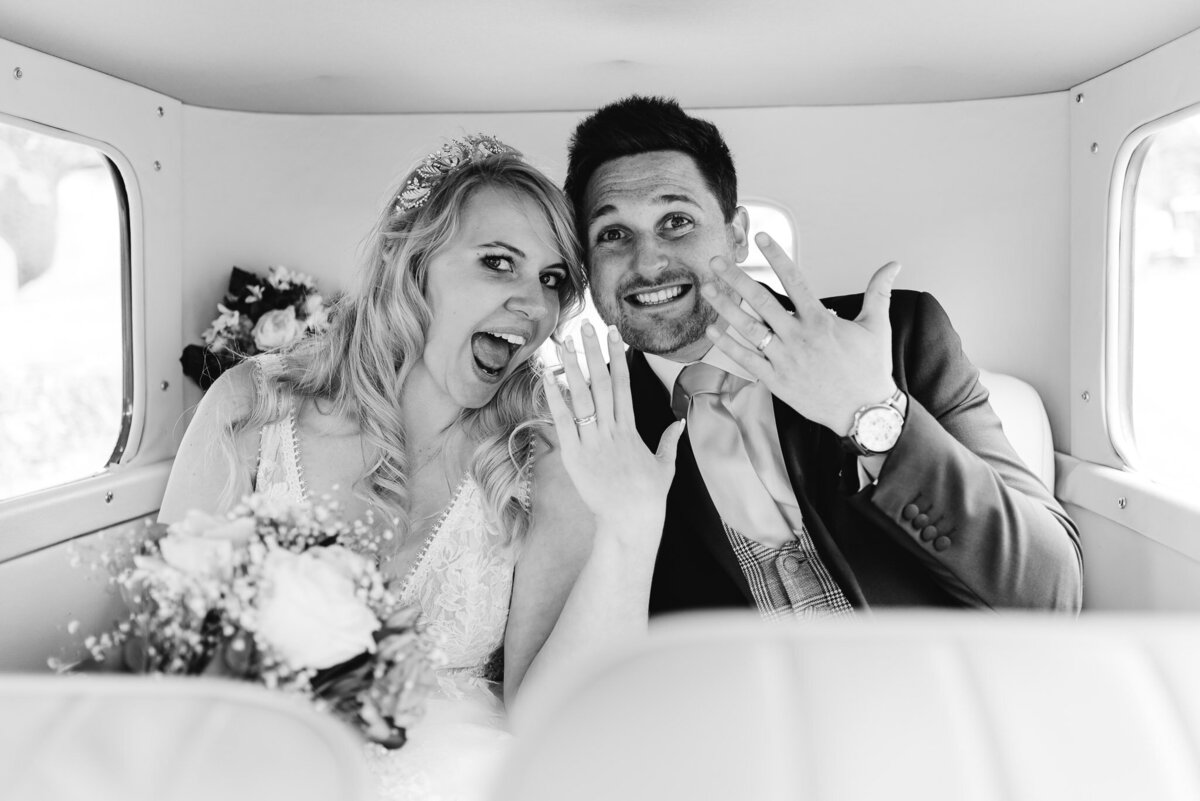 black and white image of bride and groom in the wedding vehicle smiling at the camera showing their rings