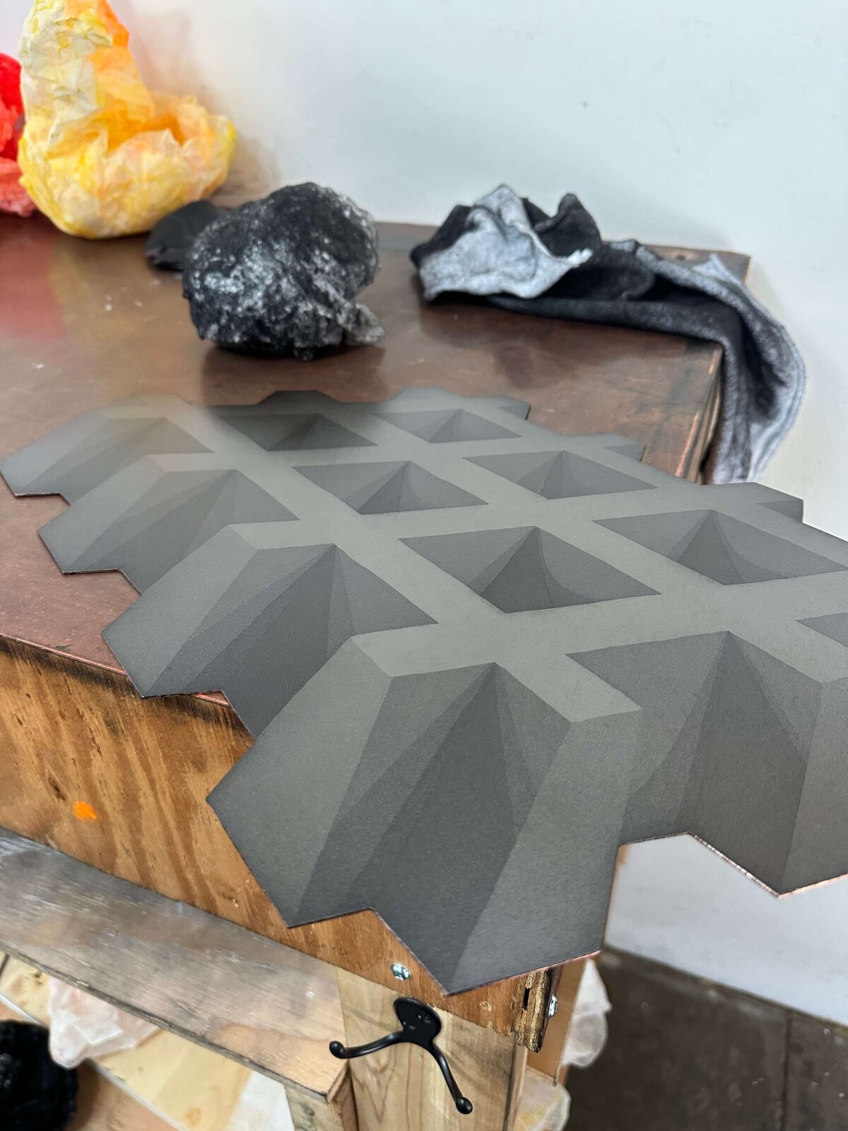 A dark, tonal aquatint print featuring a dimensional geometric form rests on a copper tabletop surrounded by stained rags.