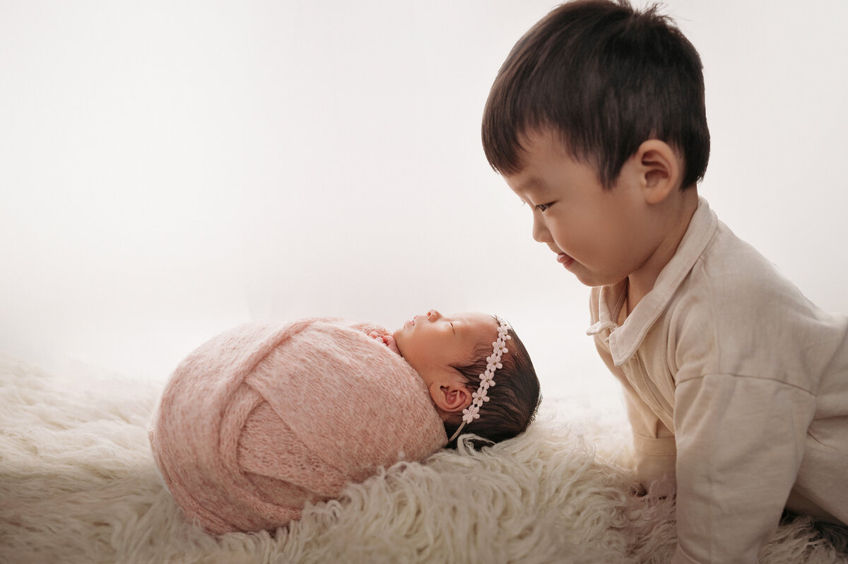 new big brother leaning over cream rug while looking at his newborn baby sister who is swaddled in a pink wrap and wearing a floral headband in front of white background
