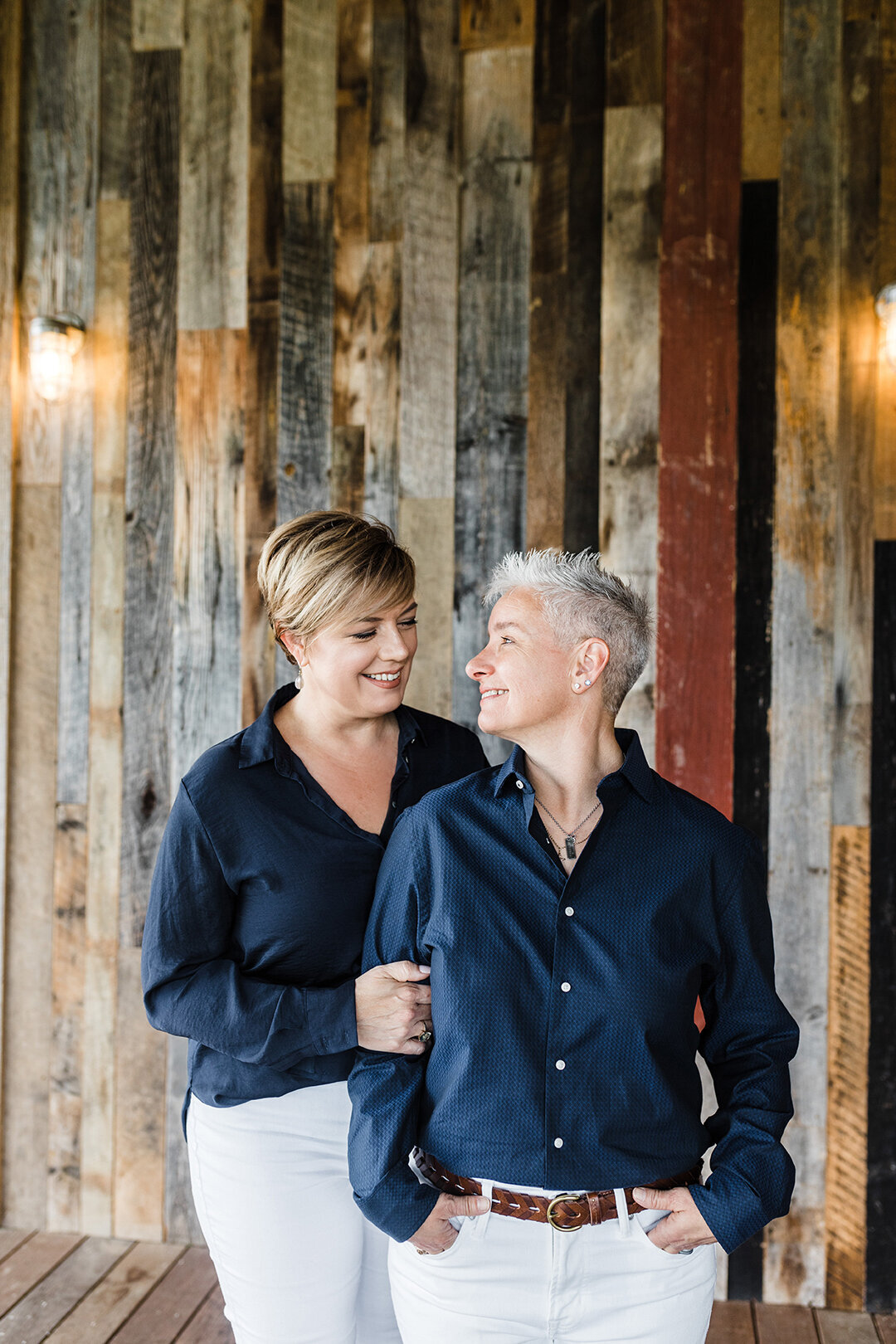 A lesbian couple standing close together and smiling at each other in front of a wood paneled wall during their engagement session in Fort Worth, Texas. They are both wearing navy blouses and white pants.