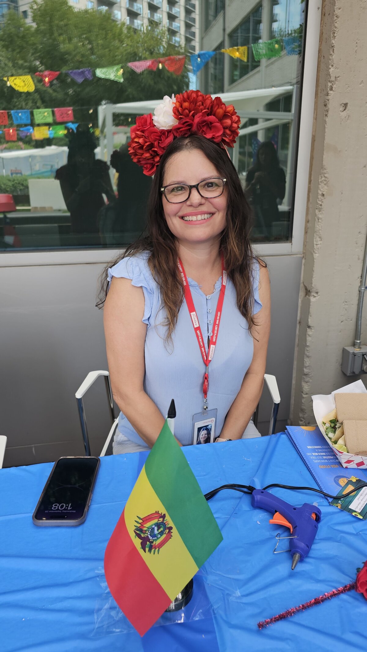 A woman in a short light blue dress, adorned with black reading glasses, proudly displays a handmade wreath of vibrant red flowers. Seated in front of a blue tablecloth table, witness the artistry come to life with some of the materials used to craft this exquisite piece.