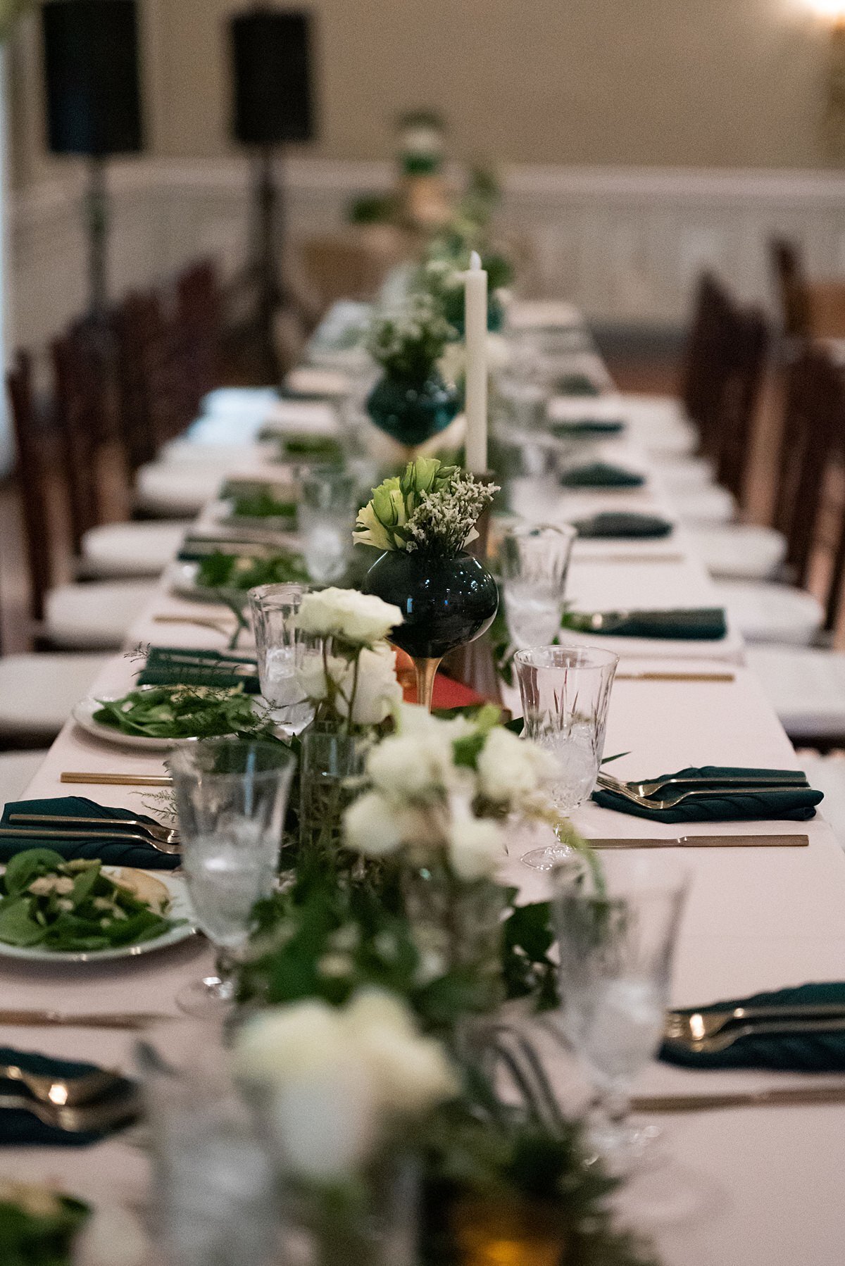 Long white table with a garland of greenery and white flowers accented with emerald and gold candle holders with white taper candles. The table is set with emerald green napkins, gold flatware and salads.