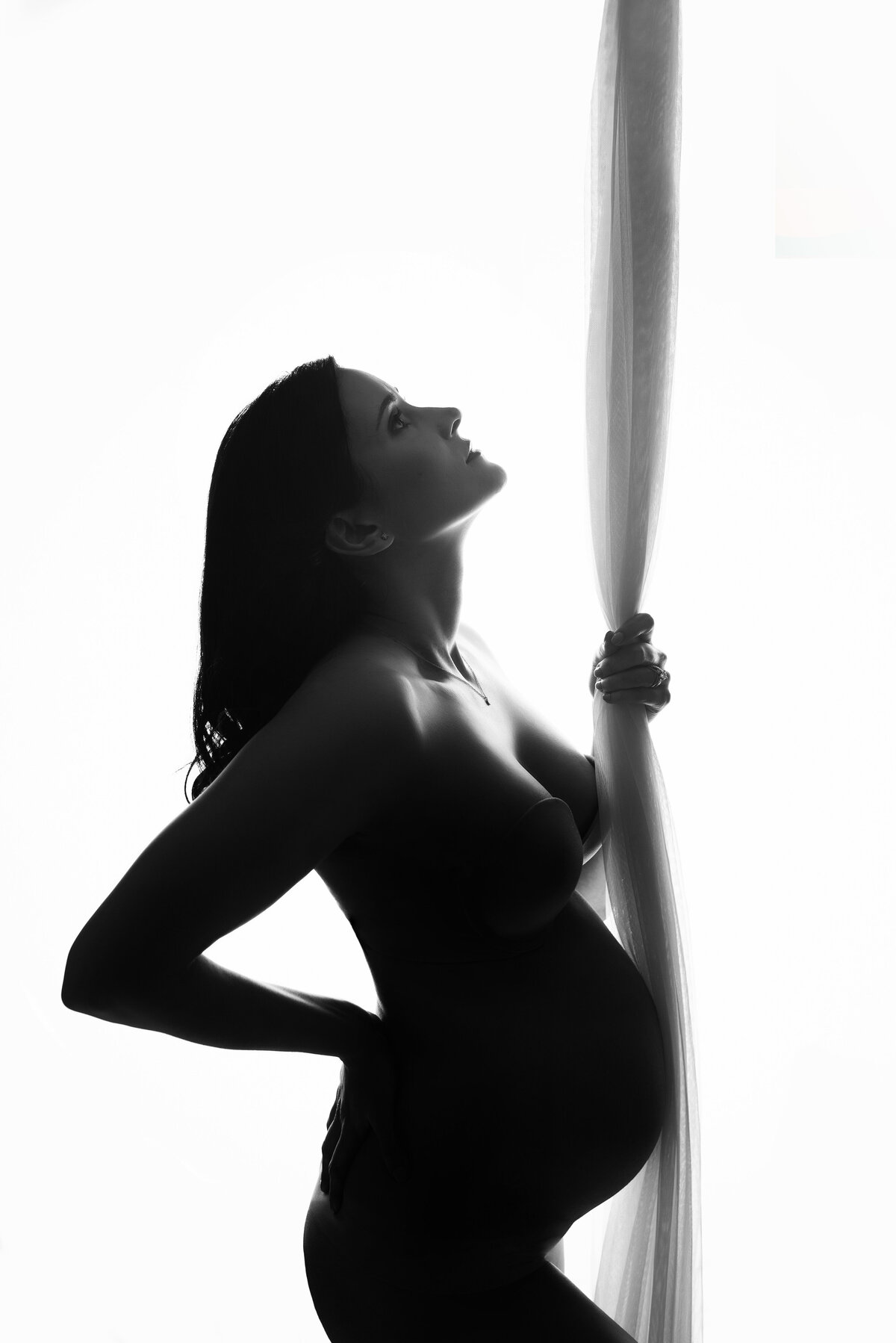 Black and white maternity portrait with woman standing to side showing her baby bump with right hand on back and left hand holding fabric coming from ceiling