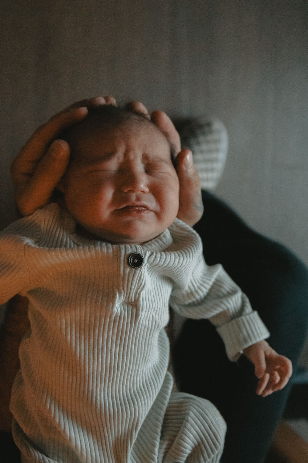 Newborn in a white onesie being gently held while crying