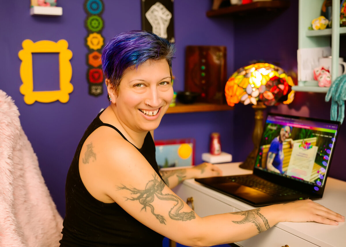 They/them podcaster sitting at their office desk in front of a laptop and smiling, while posing for a South Shore MA branding photographer