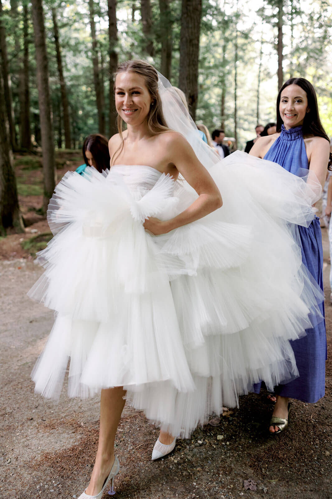 The bride is walking outdoors, lifting her wedding gown, and a bridesmaid is holding the gown's train at The Ausable Club,NY.