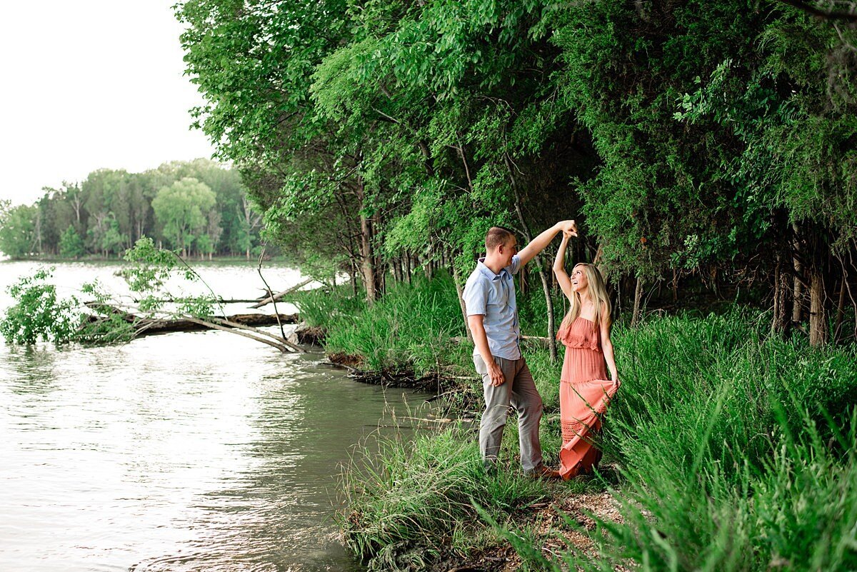 The groom twirls the bride lakeside. The groom is wearing khakis and a white button down shirt, untucked.  The bride is wearing a peach maxi sundress. The bride and groom hold their hands up as she spins around. They are standing next to a lake.