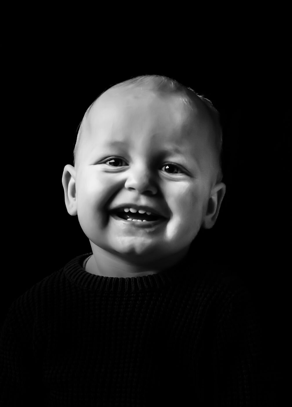Een zwart-wit portretfoto van een lachende baby, gefotografeerd tegen een donkere achtergrond.