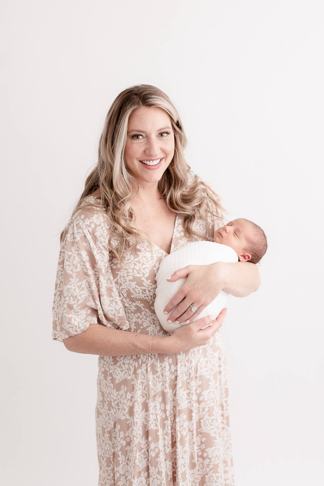 Gorgeous Mom with professional hair and makeup done and wearing a beige and white floral dress. She is holding her newborn baby who is wrapped in a white swaddle and sleeping peacefully. Mom is looking at the camera with a beautiful smile on her face.