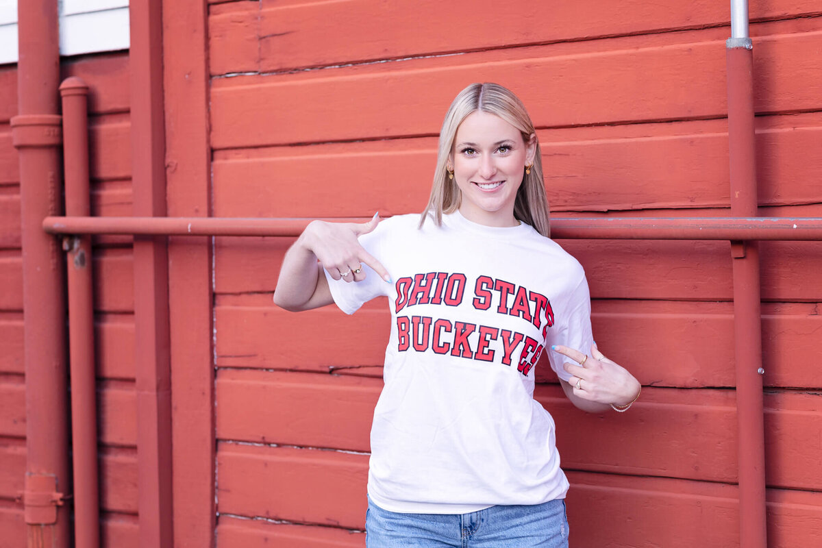A person stands in front of a red wooden wall, pointing at their white T-shirt, which has Ohio State Buckeyes in red letters. They have long hair and are smiling at the camera.
