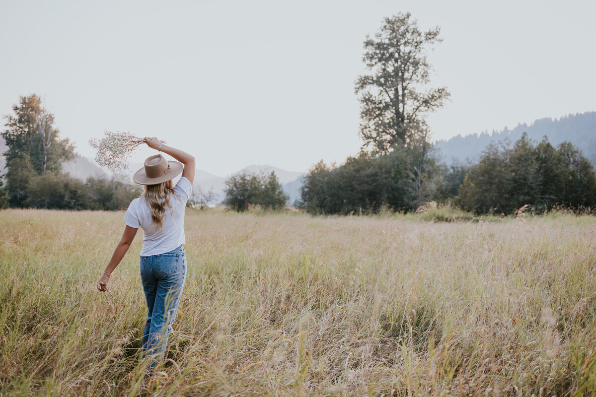 Young woman walks away from camera in grassy Idaho field while holding flowers