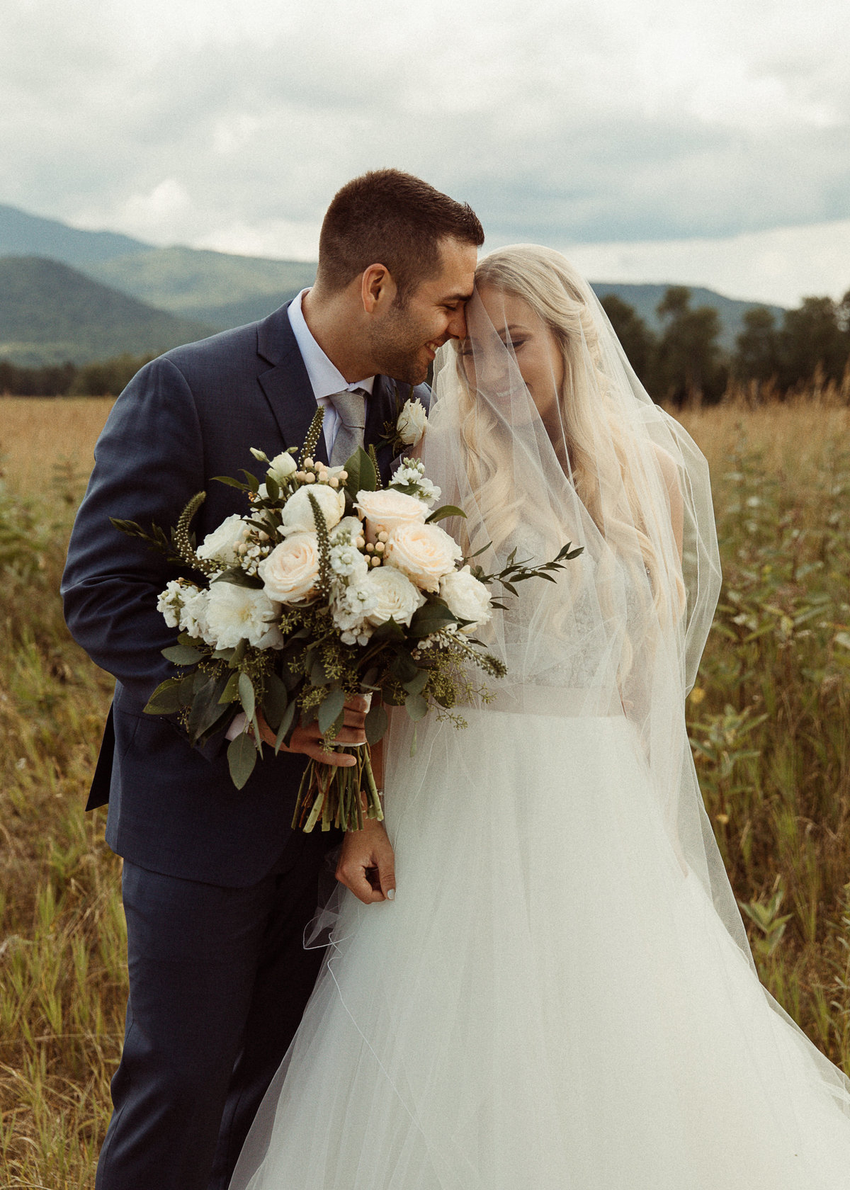Bride under her veil and groom holding a white bouquet with the Adirondack Mountains behind them in Lake Placid, NY