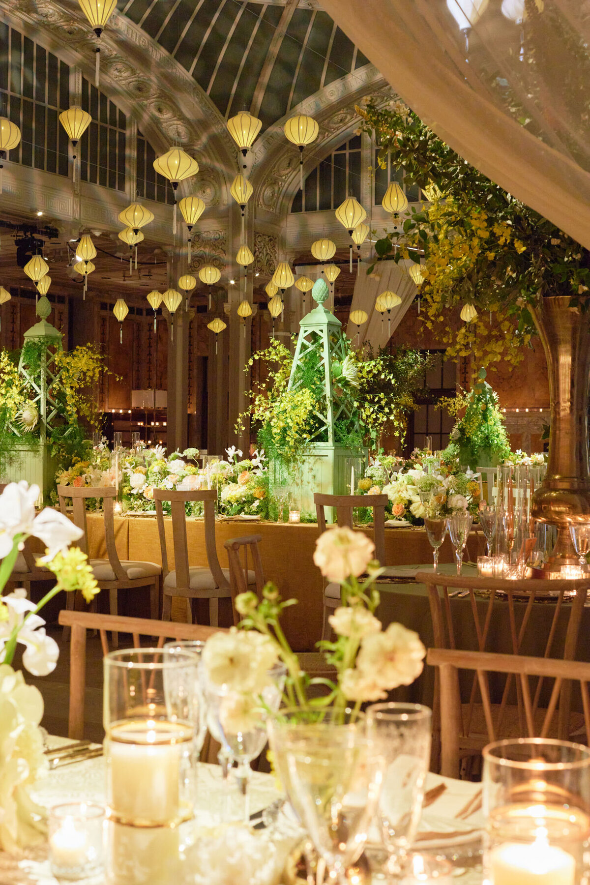 An elegantly lit greenhouse-style venue at the New York Public Library in New York City is decorated for a sophisticated event. Floral arrangements with lush greenery adorn tables set with glassware and candles. Numerous hanging lanterns illuminate the space, created by photographer Shawn Connell, creating a warm and inviting atmosphere. Aaron Navak Films is also present as the videographers capturing the special moments of the event.