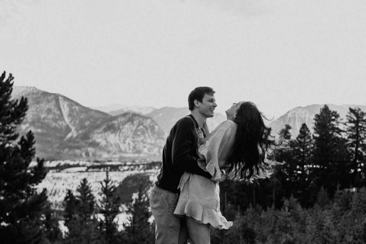 Couple laughs as he lifts her with snowy Breckenridge mountains in the distance at Sapphire Point.