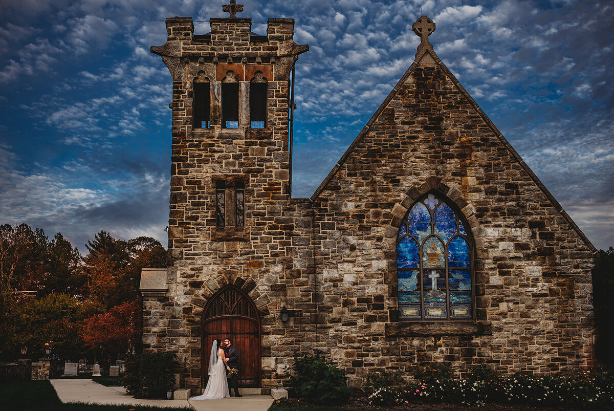 Maryland wedding photographer photographs bride and groom standing together outside of a stone chapel with large Stainglass for the Baltimore wedding