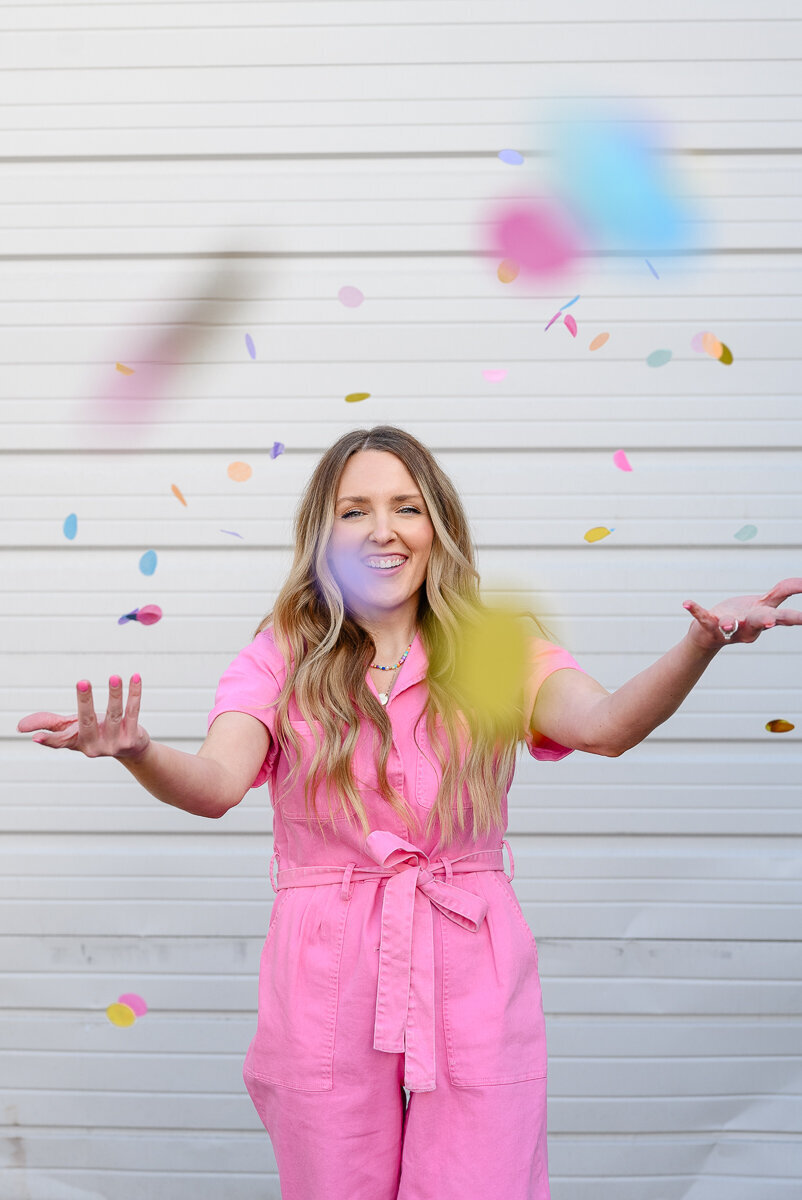 A branding photographer in Denver takes a photo of a woman in a pink jumpsuit throwing confetti to celebrate during her brand photos for her branding photography session.
