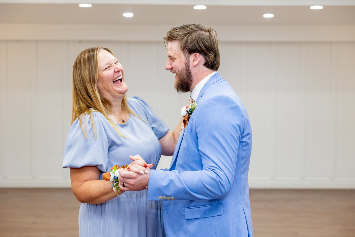The groom shares a joyful dance with his mother during the reception, both laughing and enjoying the special moment. They are wearing matching blue tones and floral boutonnieres.