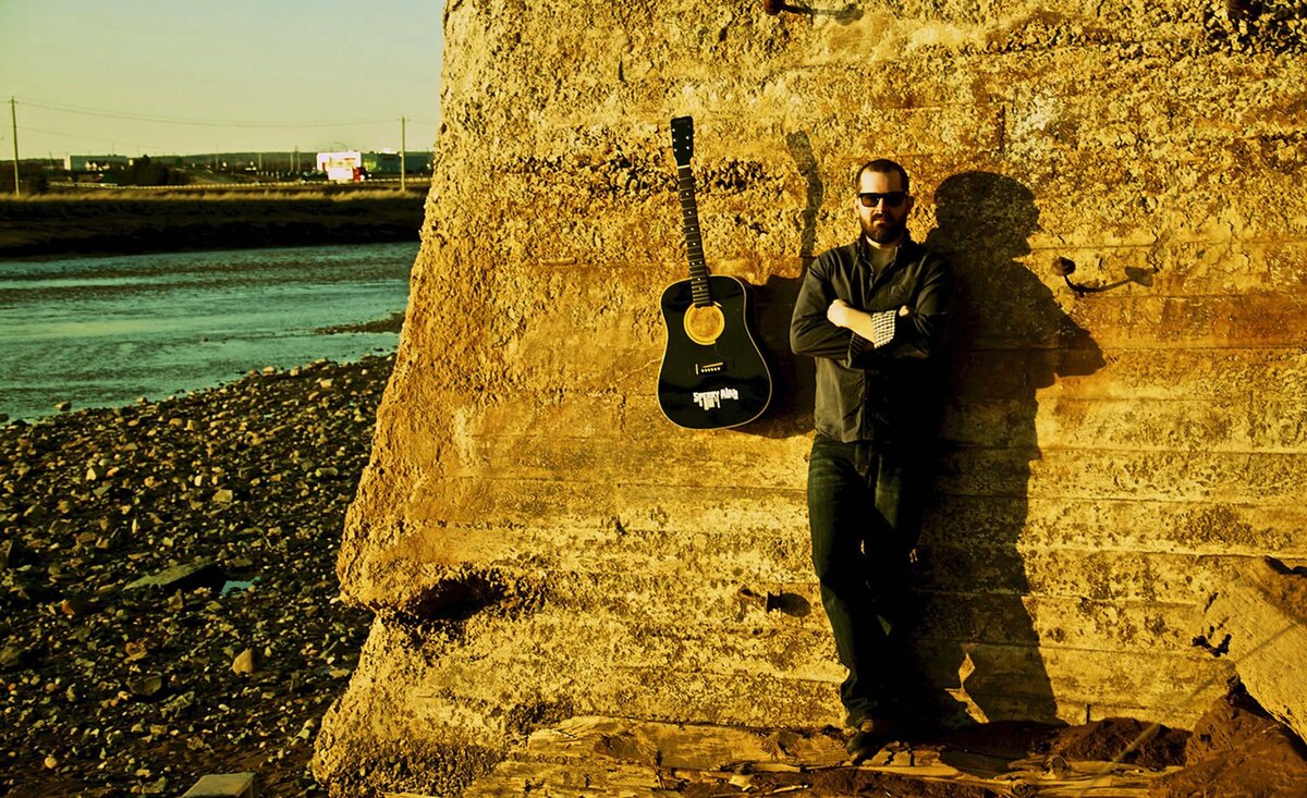 Male musician portrait Sperry Alan wearing black dress shirt black pants leaning against red rock wall black guitar hanging beside