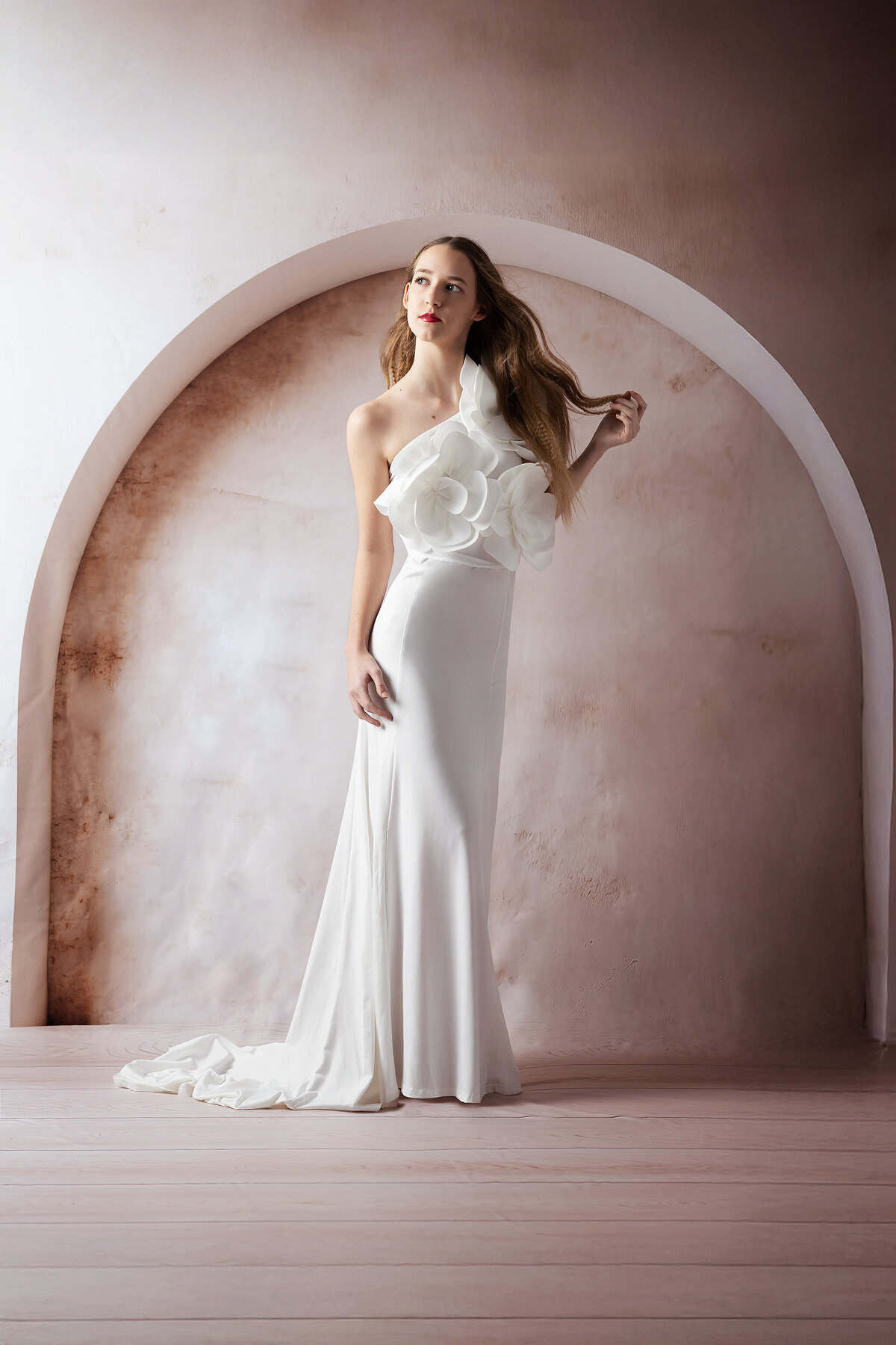 A senior in a stunning white floral gown stands gracefully against a soft pink-toned arched backdrop. The gown’s intricate floral design accentuates her elegance, perfect for a Dallas senior photographer shoot.