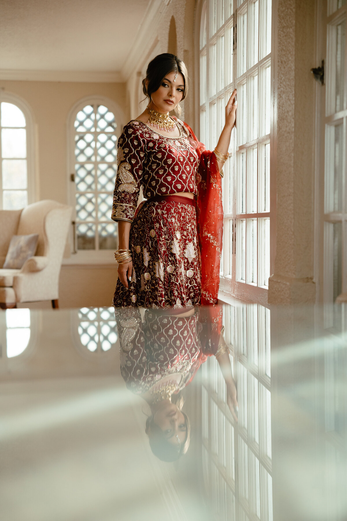 an indian bride wears a red sari and stands looking out a window while her reflection is shown in the foreground