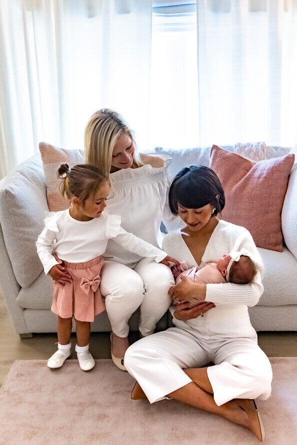 Two women and their toddler hold and look at their newborn on the couch at their house.