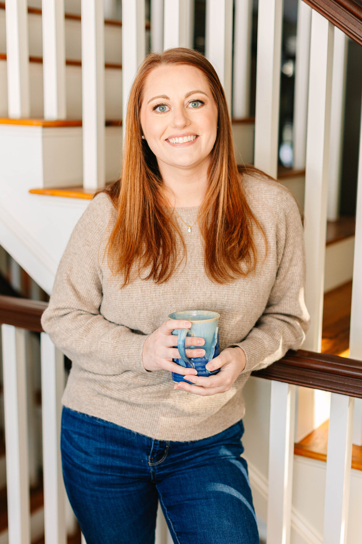 A woman with long red hair and green eyes smiles while leaning against a staircase railing, holding a blue ceramic mug. She wears a beige sweater and dark jeans, with warm sunlight illuminating the scene