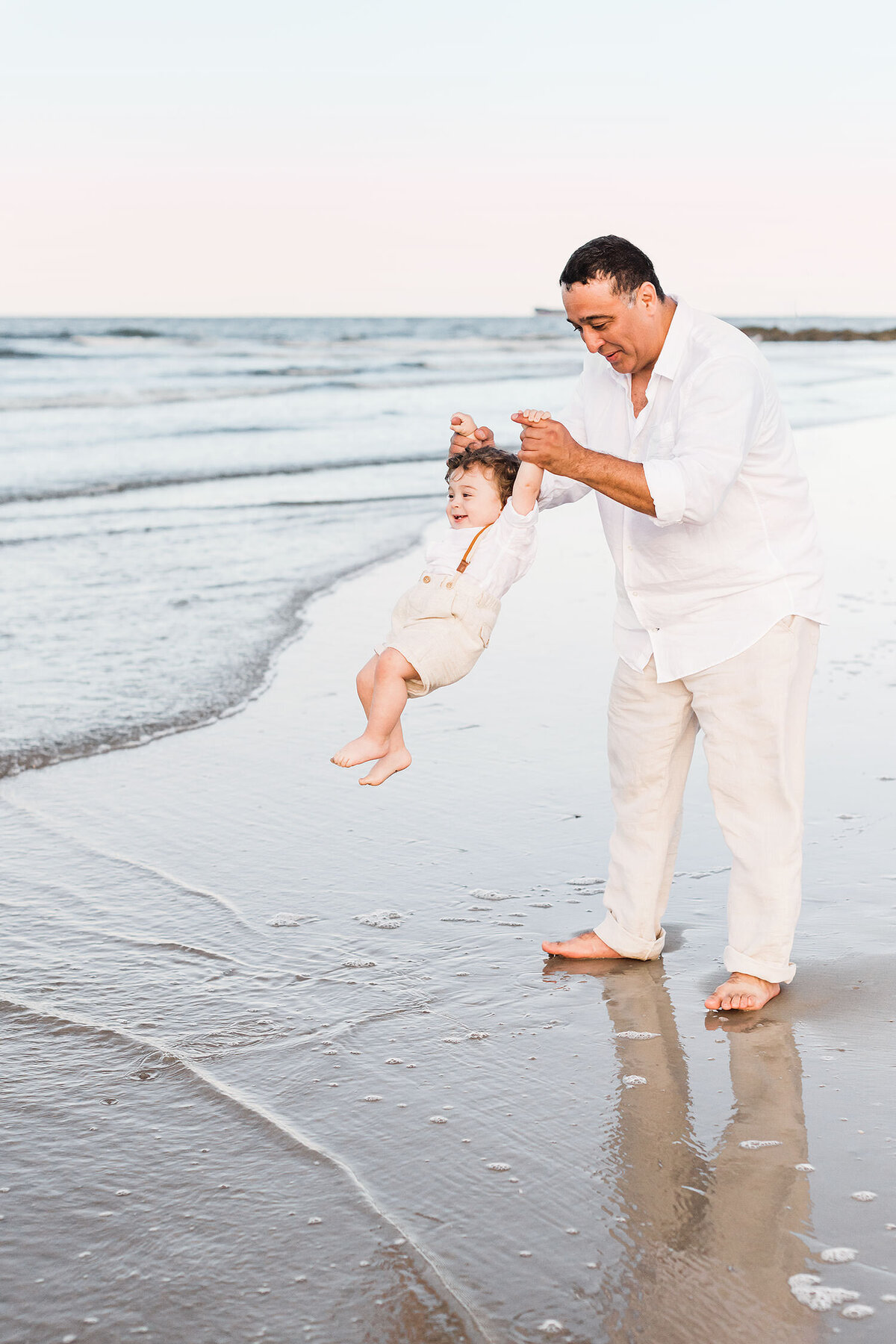 a father dressed in white is swinning around his toddler near the ocean