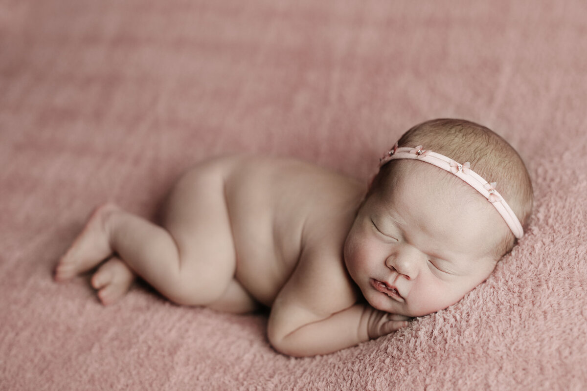 Studio newborn photography - Baby girl curled up sleeping on her side. Baby is bare wearing a petite pink headband. Her hands are resting under her chin.