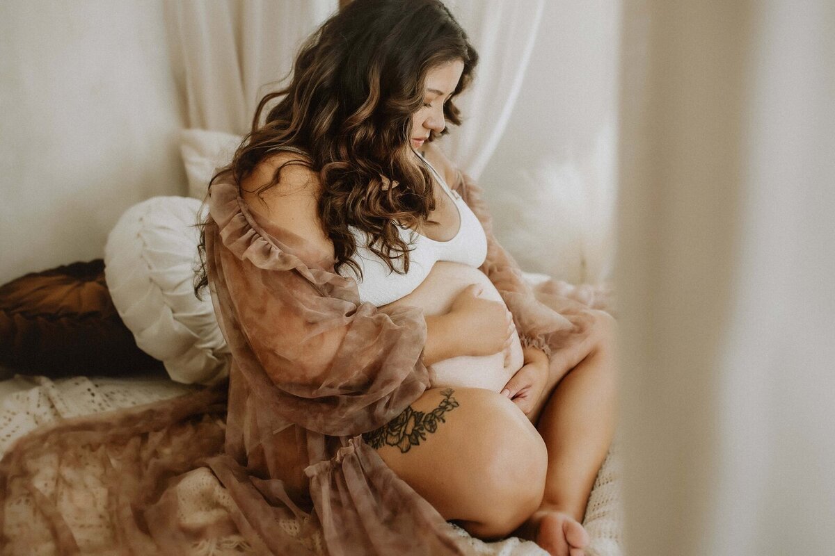 A pregnant woman sits on a bed, draped in a sheer robe and white top, cradling her belly with both hands. The decor features white and brown pillows, capturing the serene moment beautifully—an intimate scene worthy of a Macon studio maternity photographer's lens.