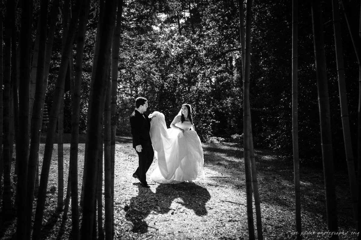 A groom holding a bride's train as they walk in a wooded area.