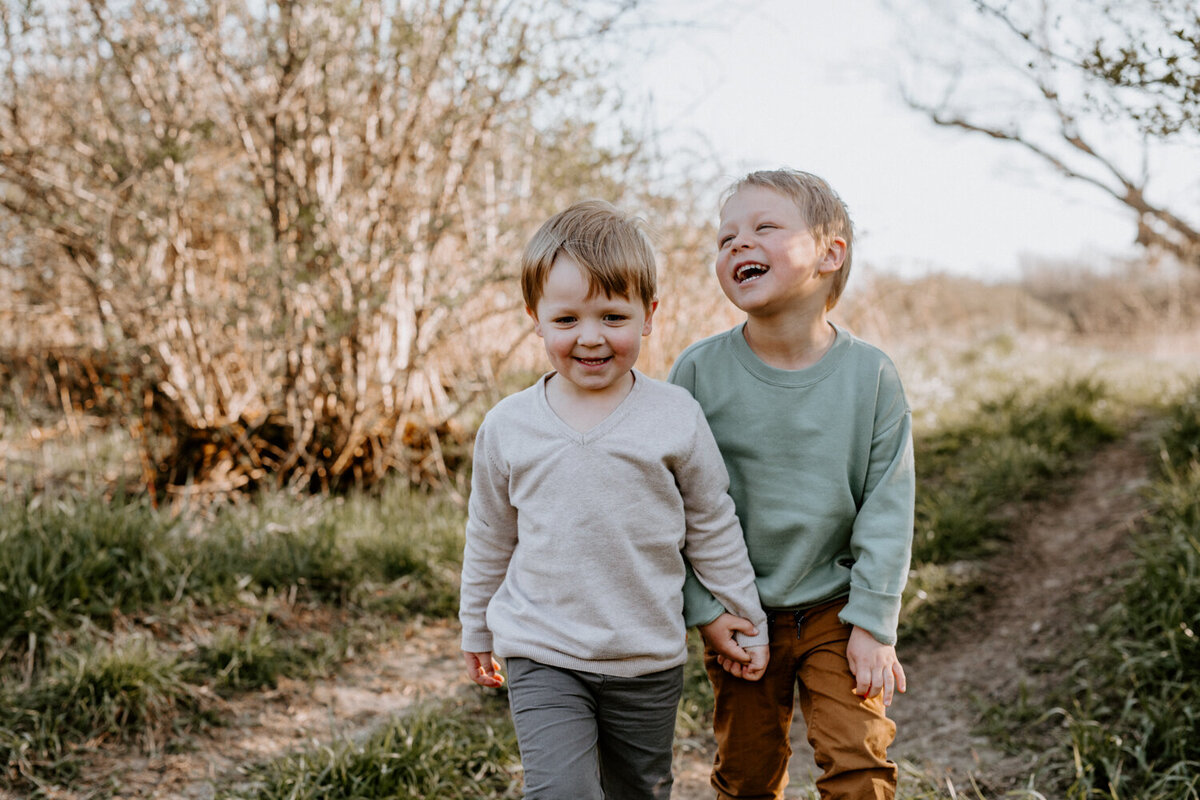 Two young brothers are holding hands walking down a trail for a top family photographer in Exeter, Ontario. The brother are laughing, one toward the camera, the other off in the distance.