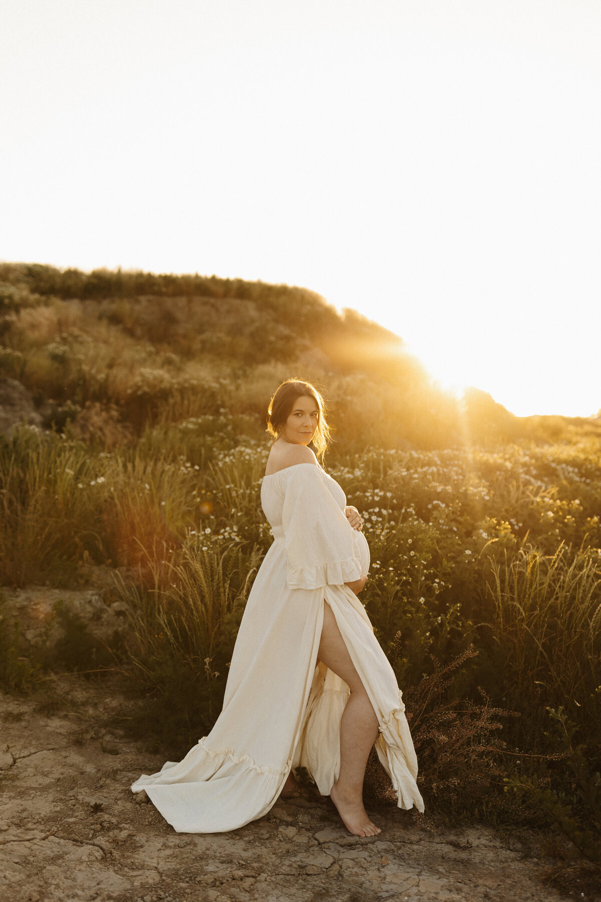 a woman with brown hair stands wearing a white gown during a maternity photoshoot and poses for the camera holding her pregnant belly while she's photographed by christchurch photographer lucy jane