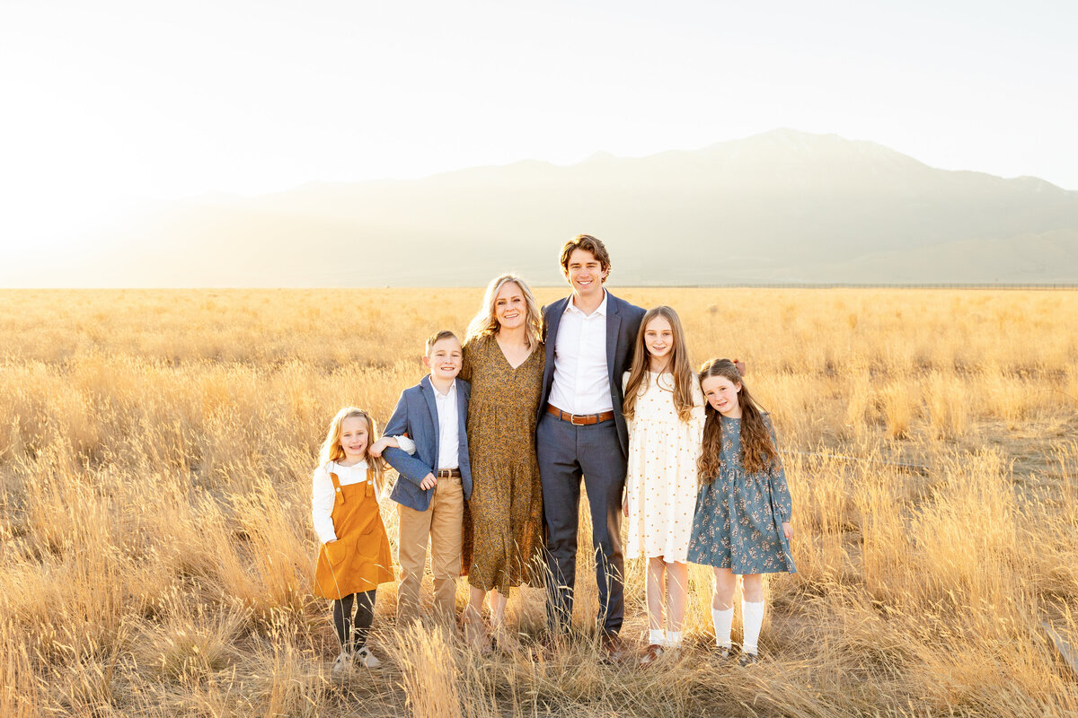  A beautiful family of six poses together in a sunlit field, dressed in coordinated autumn colors with mountains in the background.