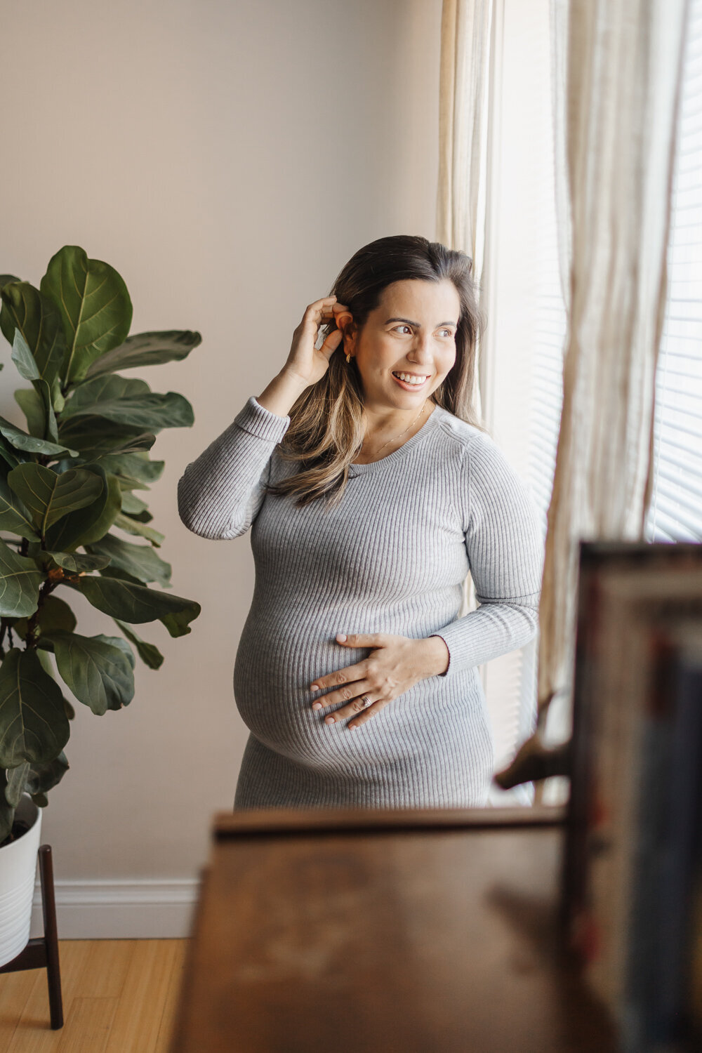Pregnant woman in grey dress posing next to bedroom window