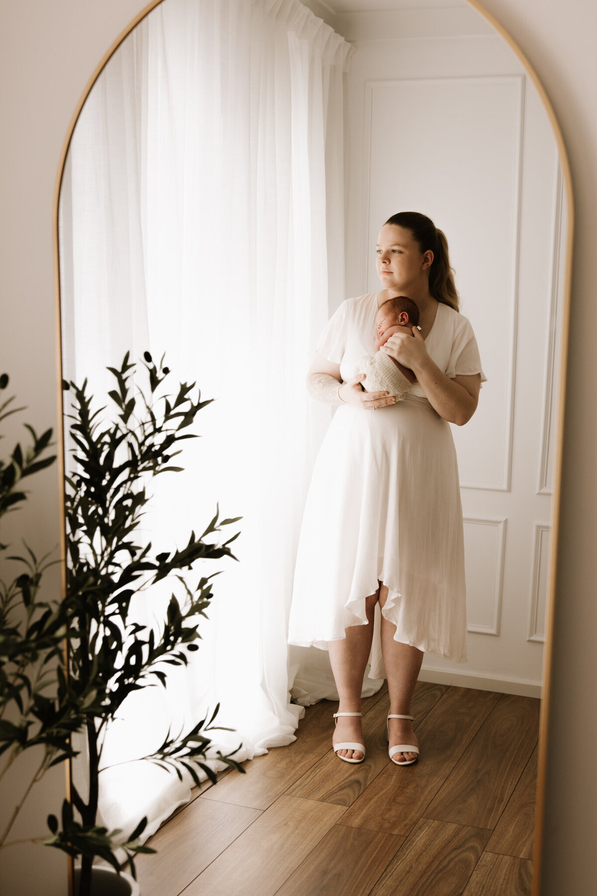 A mother stands in front of soft sheer curtains in a warm, light-filled studio, cradling her newborn close to her chest. She wears a flowing white dress, and her baby is wrapped in a cream knitted outfit. The moment is captured through the reflection of an arched mirror, with a potted plant in the foreground adding a natural touch. The atmosphere is calm, elegant, and intimate.