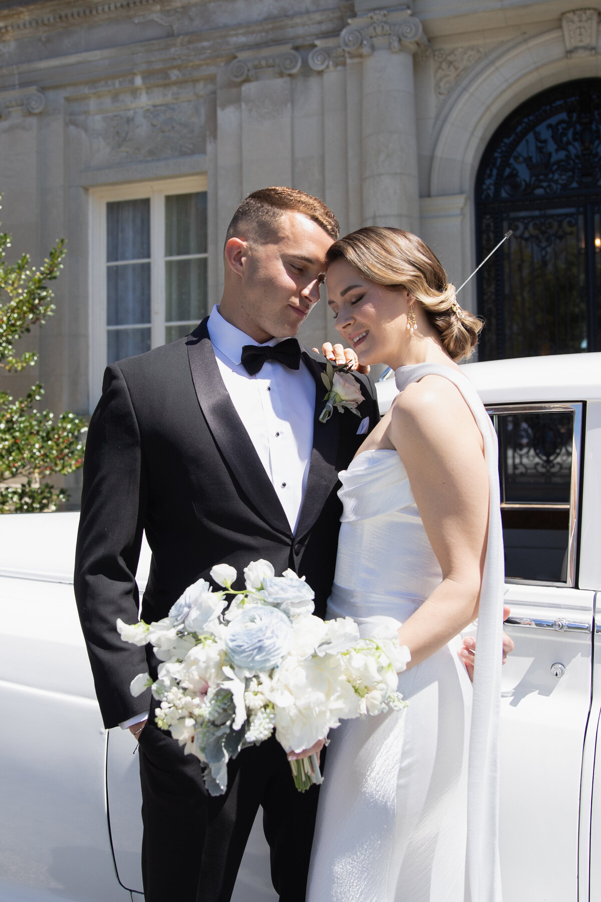 Aldrich Mansion Wedding Photos | Kelsey Sheehan Photography Timeless Rhode Island Weddings | A couple in formal wedding attire stands lovingly beside a classic white car. The bride holds a bouquet of white flowers, conveying elegance and joy.