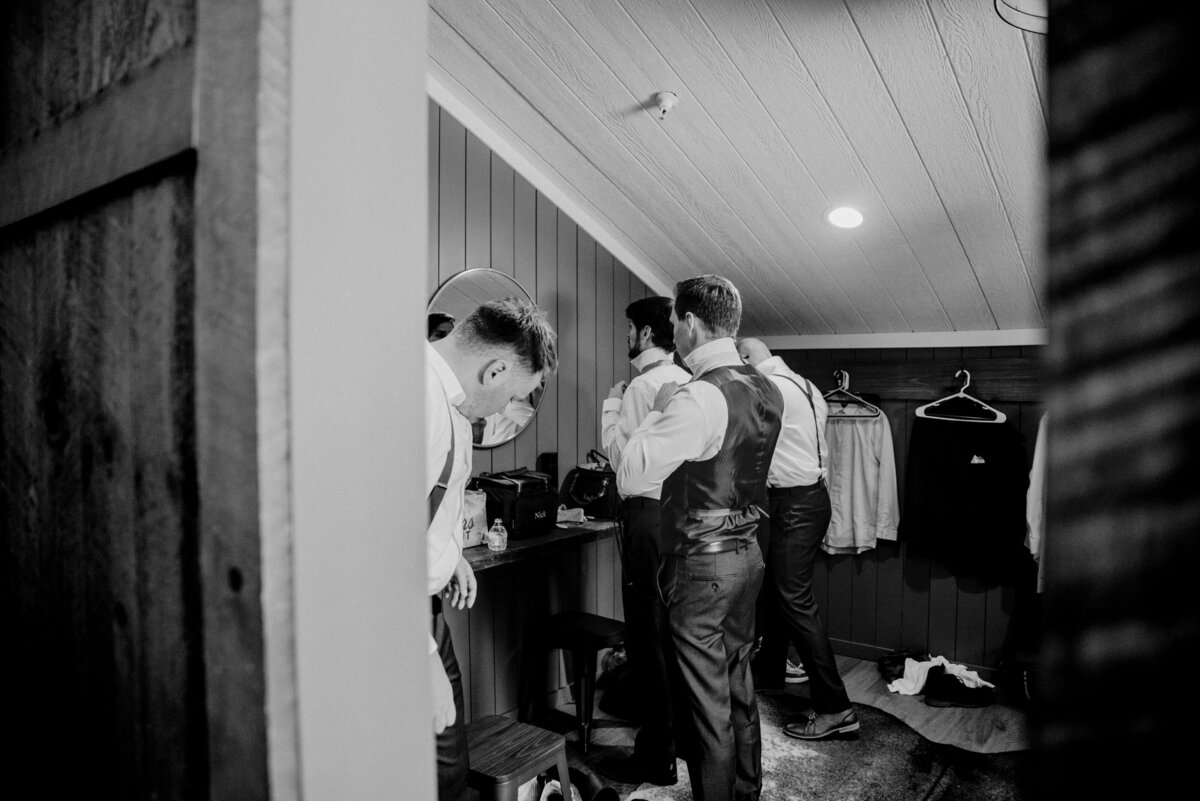 Black and white glimpse into a moment of preparation: a gentleman, fully engrossed, adjusts his bow tie in front of a mirror, while others nearby attend to their own attire, against the backdrop of a narrow, elegantly paneled room taken by jen Jarmuzek photography a Minneapolis wedding photographer