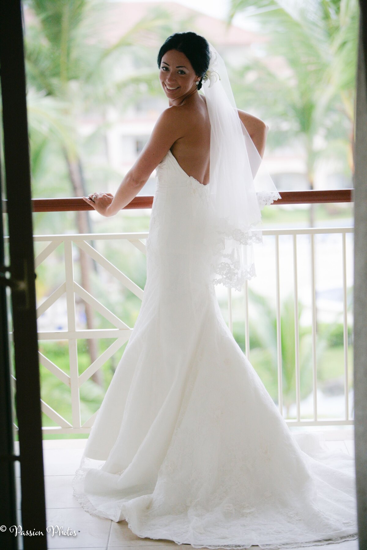 A bride stands on a balcony, looking over her shoulder and smiling with joy. This captivating image captures the elegance and excitement of destination weddings, highlighting the beautiful and memorable moments we specialize in showcasing through our wedding photography.