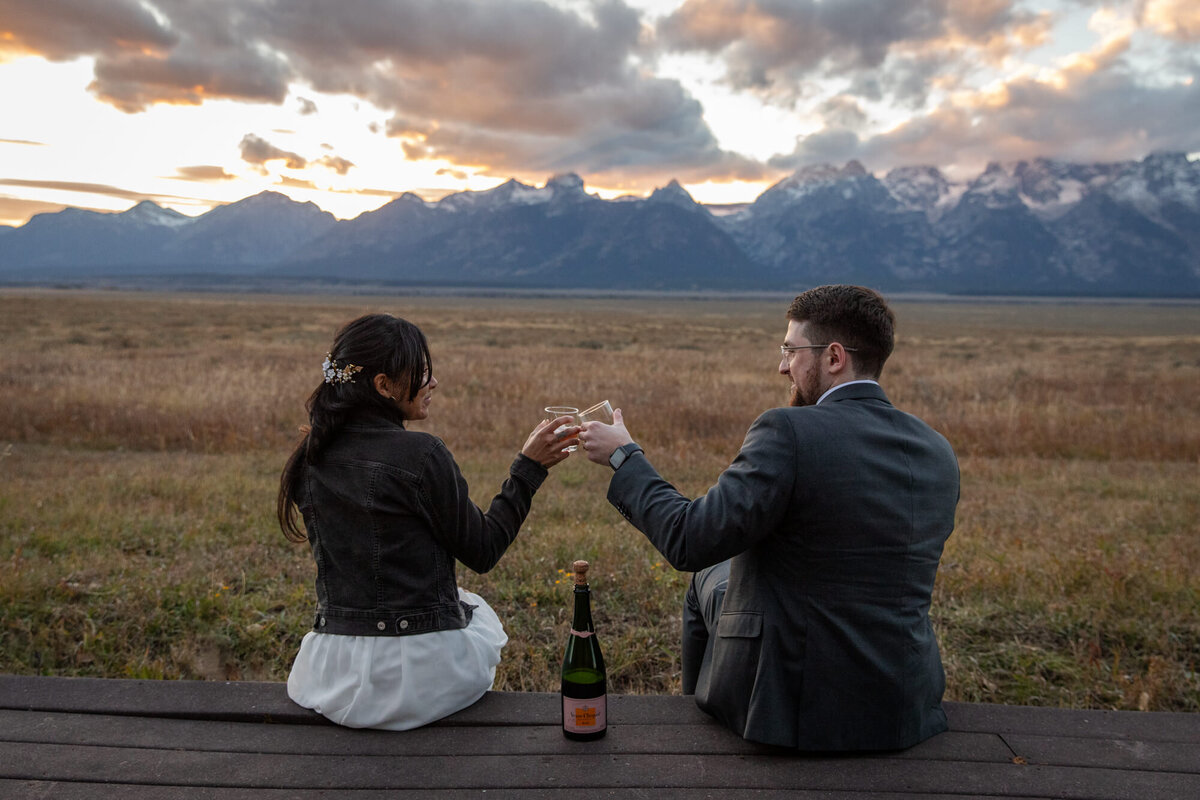 A bride and groom sit on a wooden deck, toasting their glasses as the sunsets around them.