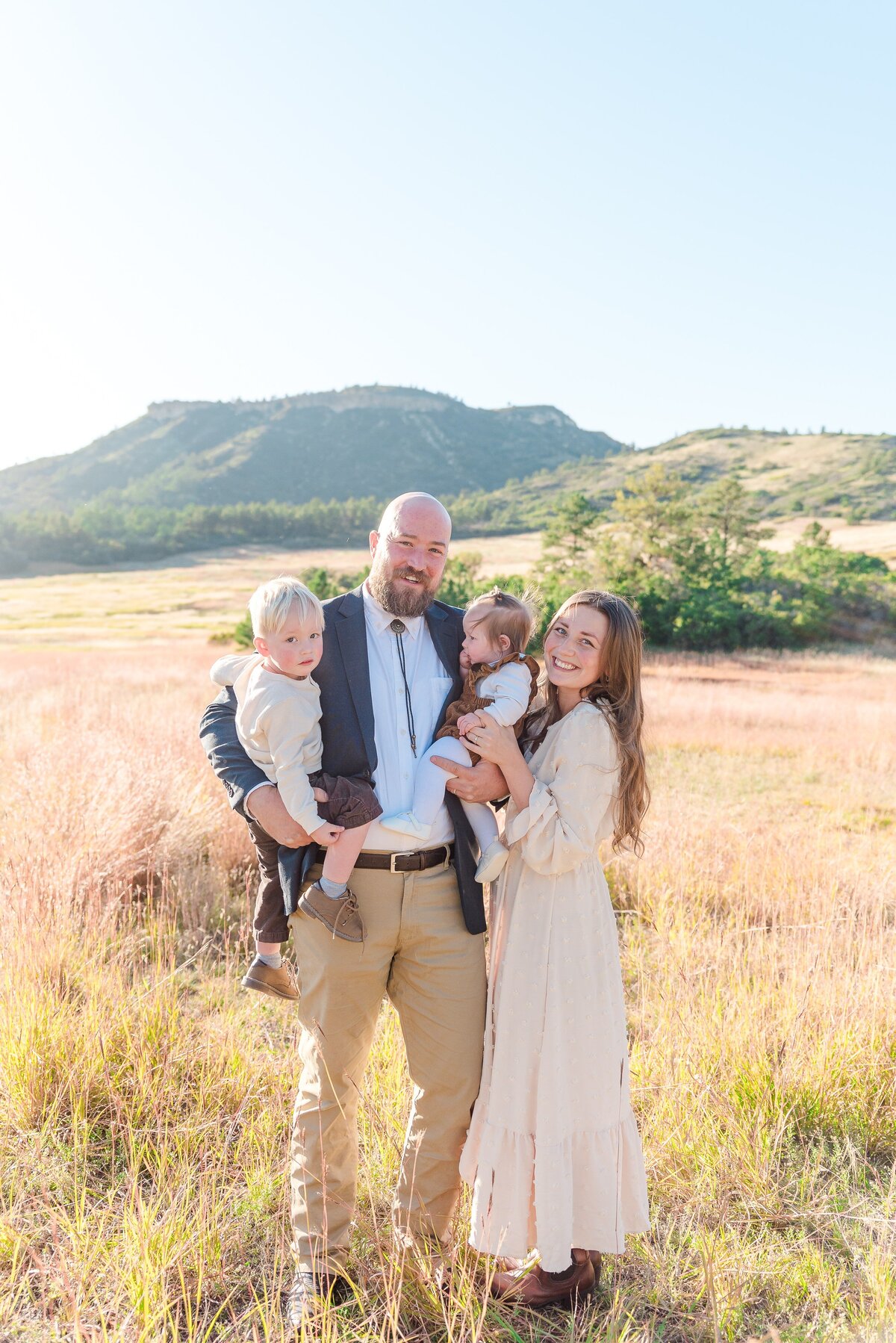 a family with a mom, dad, infant daughter, and toddler son stand close together while mom and dad hold the kids and smile for the denver family photographer