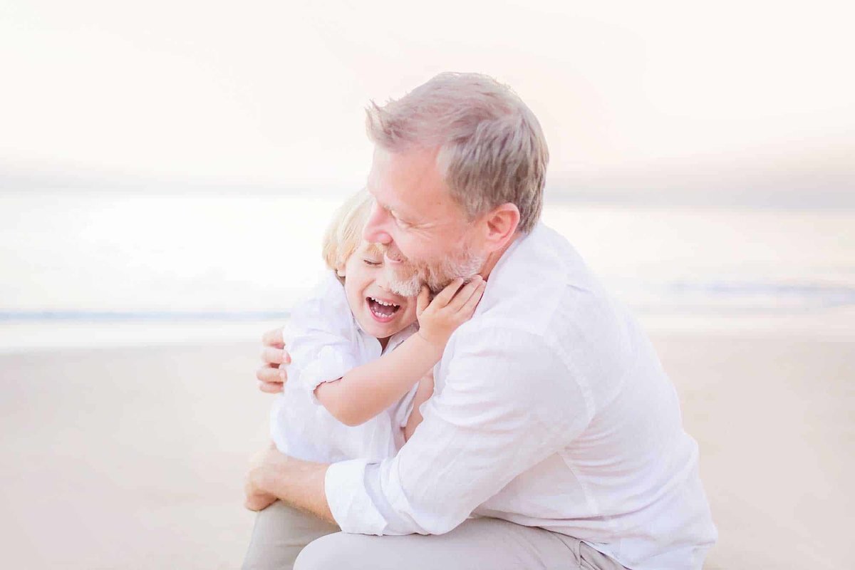 Dad snuggles his young son on the beach in Maui at sunrise as Love + Water captures the young boy laughing on the sand